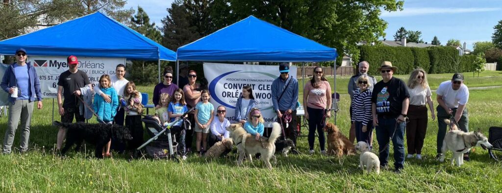 A group photo of participants (people and dogs) at the 2024 Dog Walkathon. They are standing in front of two blue tents with banners for Convent Glen Orleans Wood Community Association and for sponsors Myers Orleans dealerships. There are about 20 people in the photo. Une photo de groupe des participants (personnes et chiens) au 2024 Dog Walkathon. Ils se tiennent devant deux tentes bleues avec des bannières pour Convent Glen Orleans Wood Community Association et pour les sponsors Myers Orleans dealerships. Il y a environ 20 personnes sur la photo.
