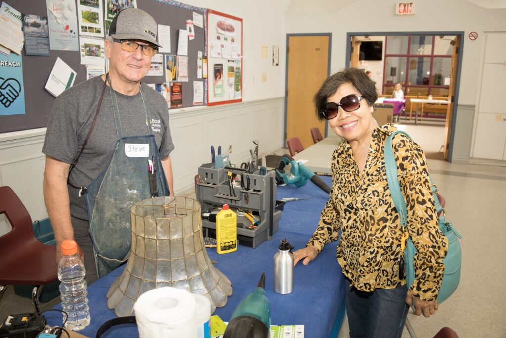 photograph of a woman at a repair station. She and the volunteer are posing with a lamp shade.